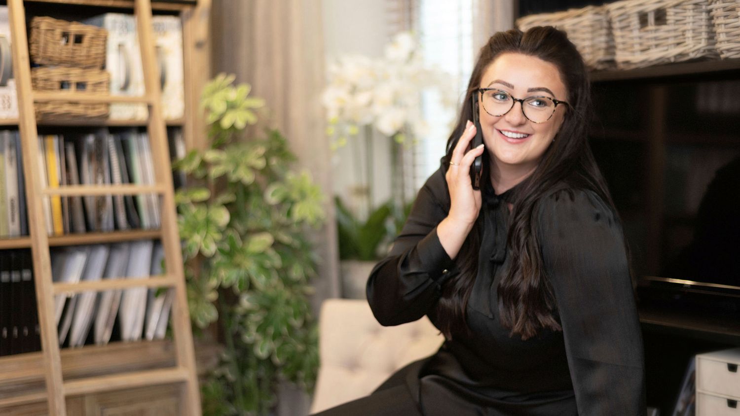 Young woman with glasses smiling while talking on phone in stylish home office setting.