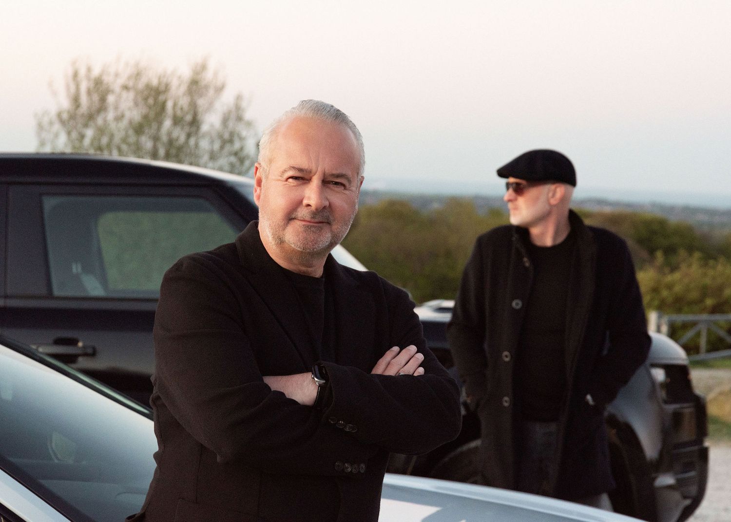 Two men in black clothing posing near a dark car outdoors at dusk with scenic countryside in the background.