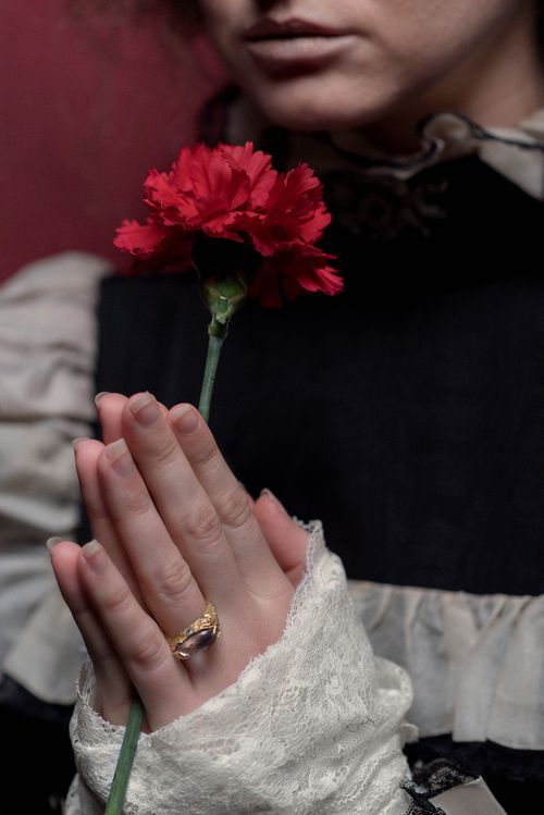 Person in vintage attire holding a red carnation flower with a gold ring on their finger.
