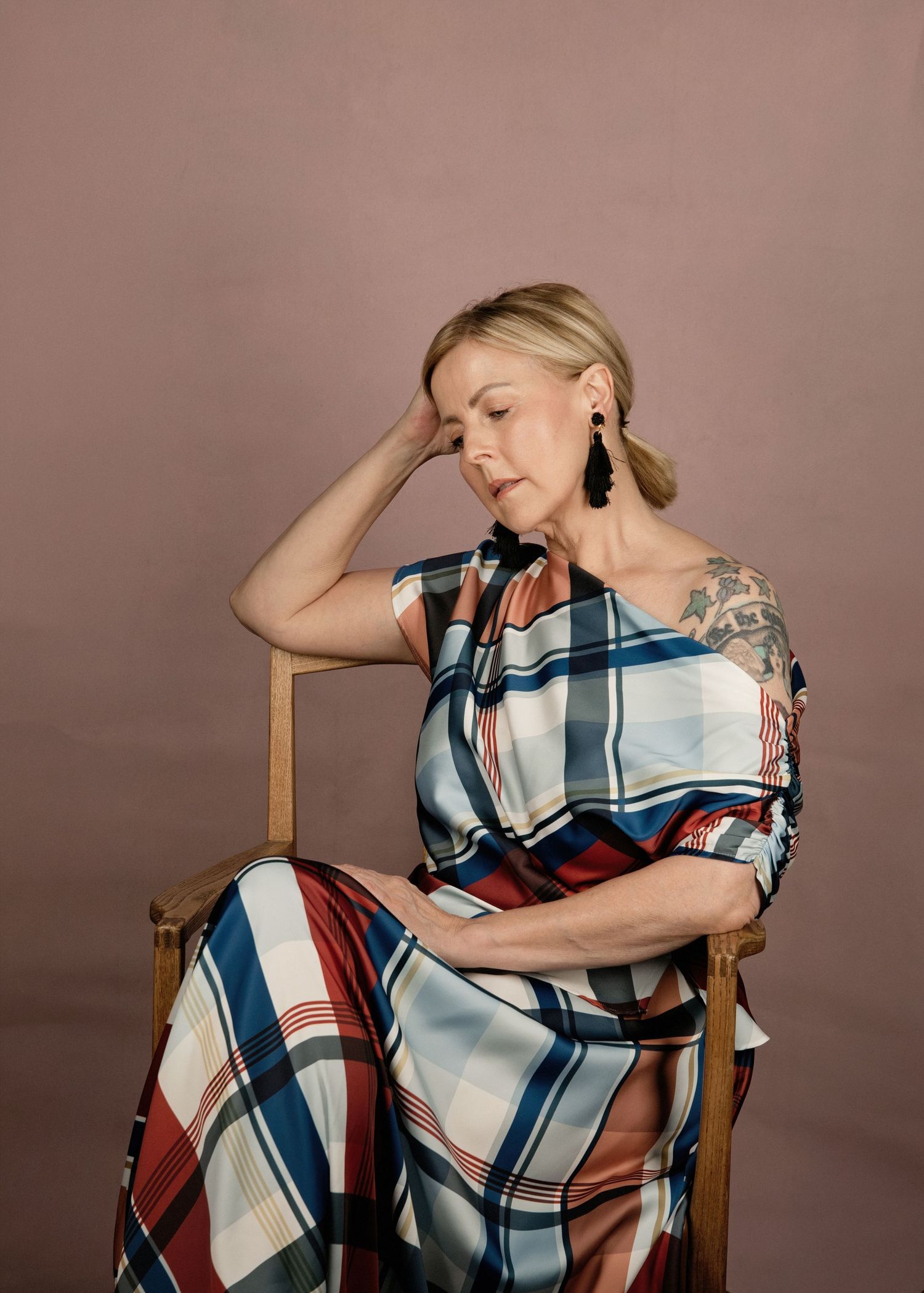 Woman in plaid dress seated on wooden chair, hand raised to head, wearing black earrings, against pink background.