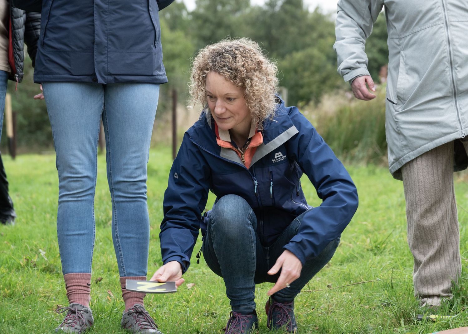People in navy jackets and jeans kneeling in grass examining something on the ground in a line formation.