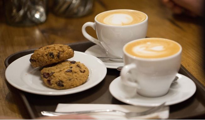Two cappuccinos with latte art served alongside chocolate chip cookies on white plates.