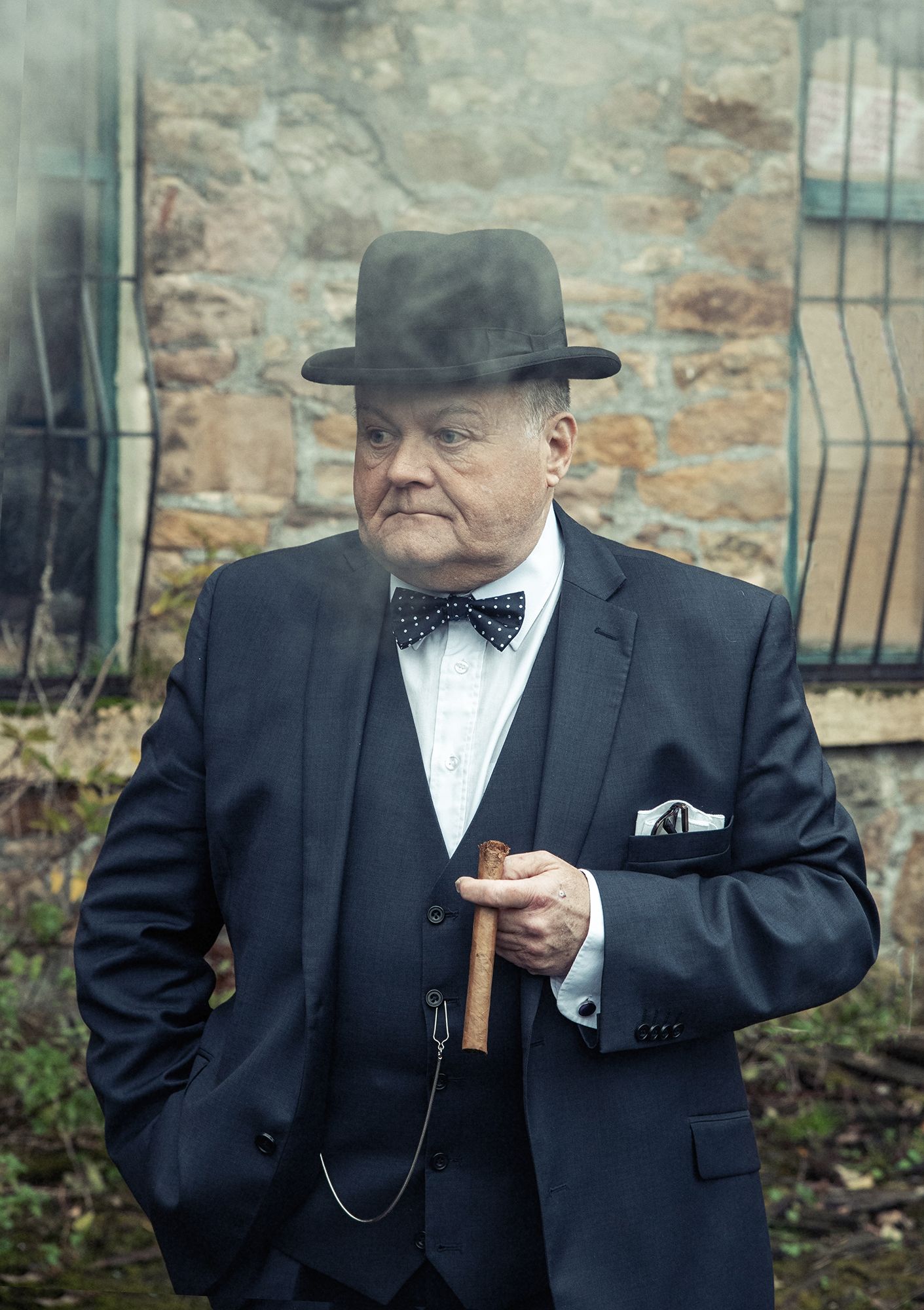 Gentleman in formal attire with bow tie and bowler hat poses against stone wall.