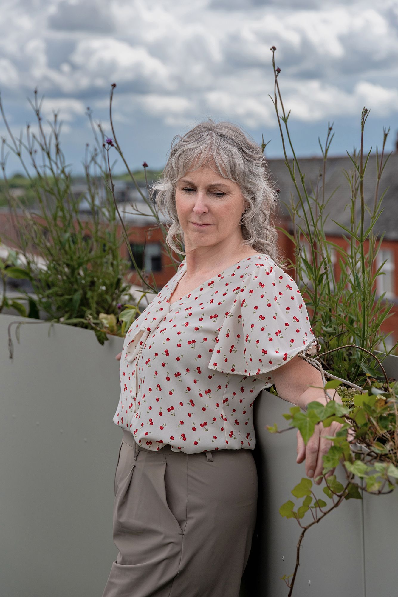 A middle-aged woman with gray hair in a polka dot blouse stands by rooftop planters under a cloudy sky.