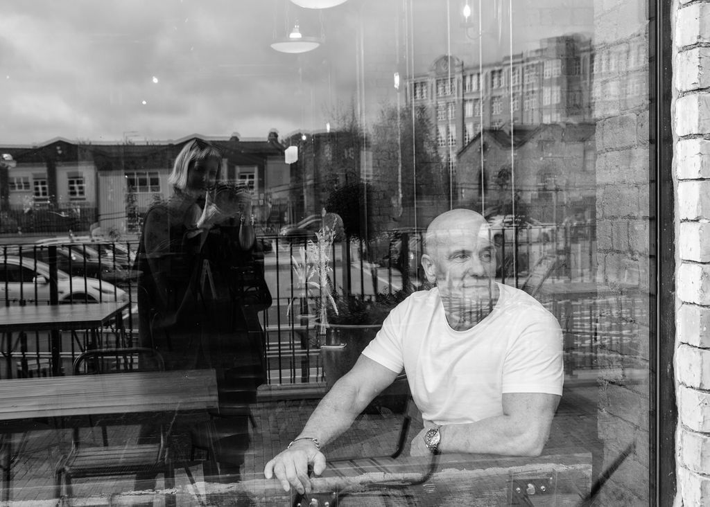 Black and white photo of a man in white t-shirt sitting at a table, with a woman's reflection in the window behind him.