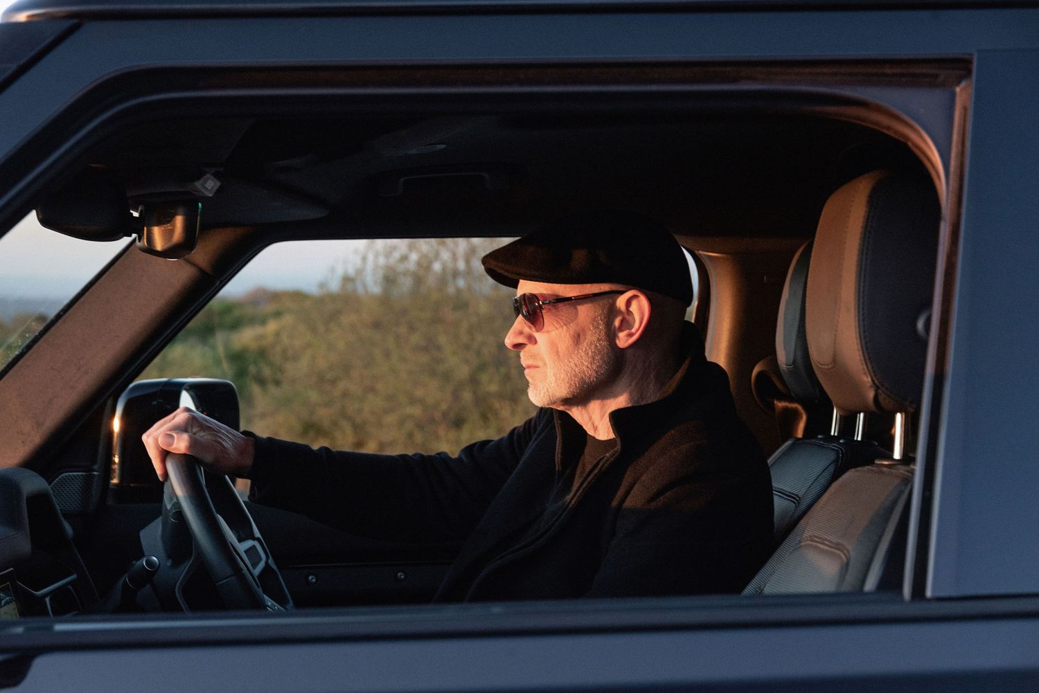Man wearing sunglasses and cap driving an SUV through scenic countryside at golden hour sunset.