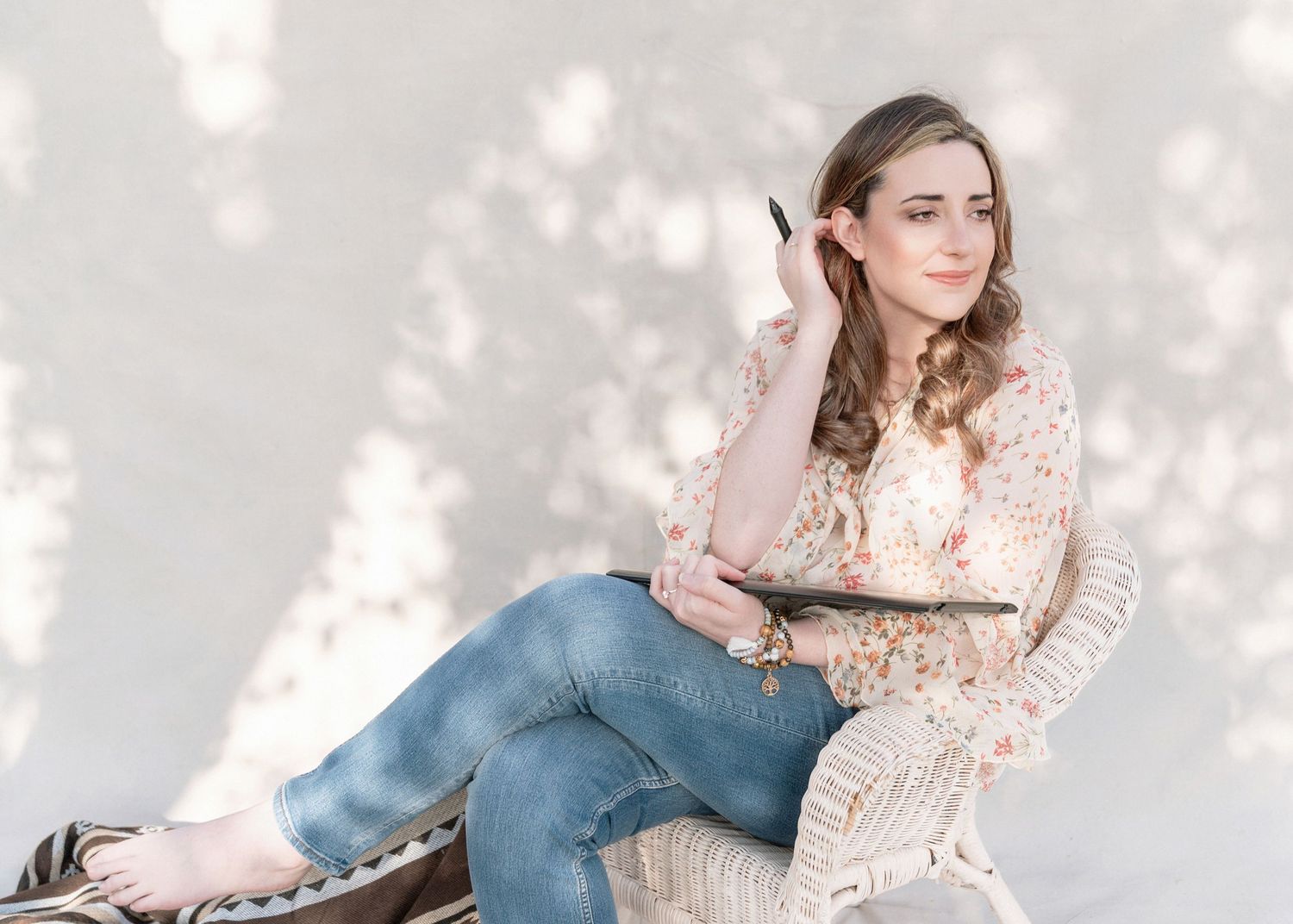 Young person in lace top and jeans relaxes on wicker chair in soft natural lighting.