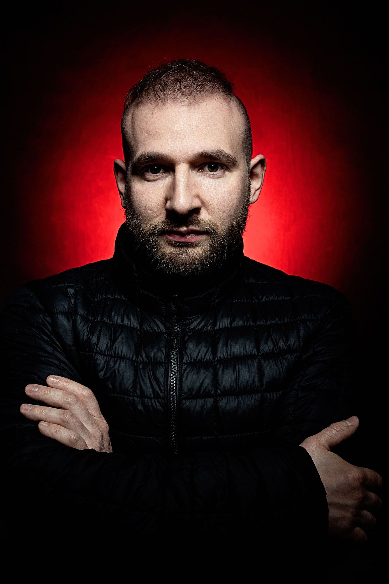 Dramatic portrait against red background of person in black puffer jacket with arms crossed.