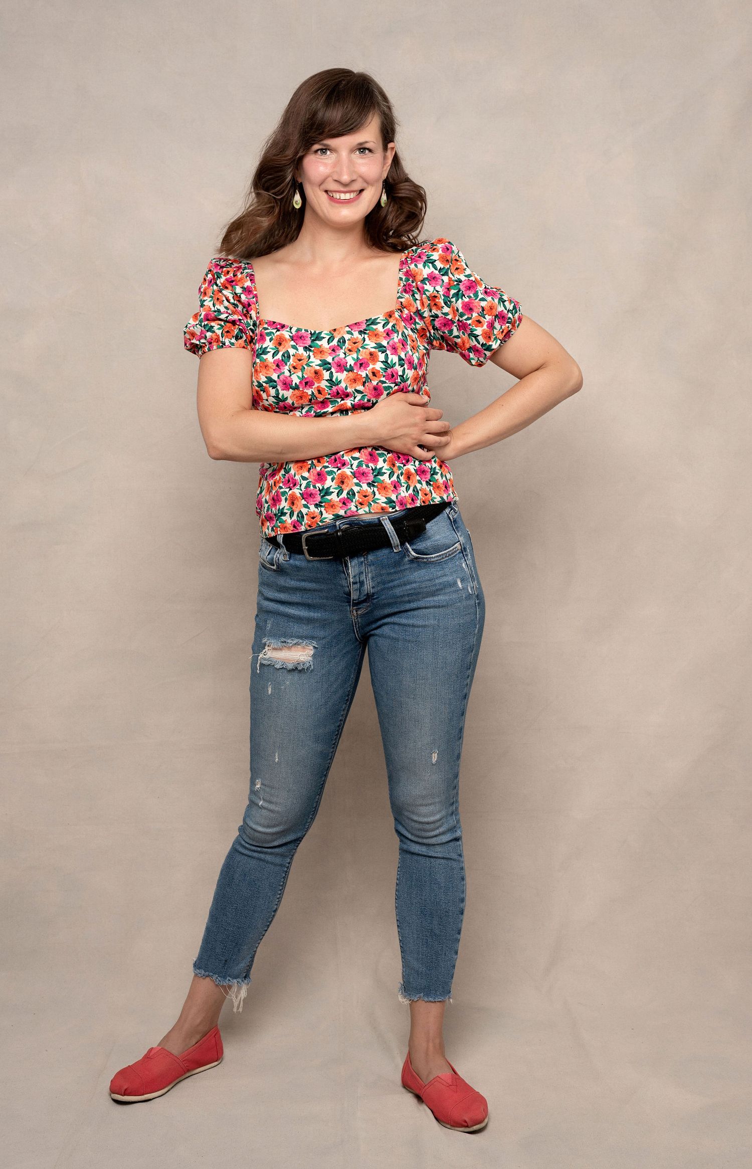 Full-length casual portrait in floral top, distressed jeans and red flats against neutral background.