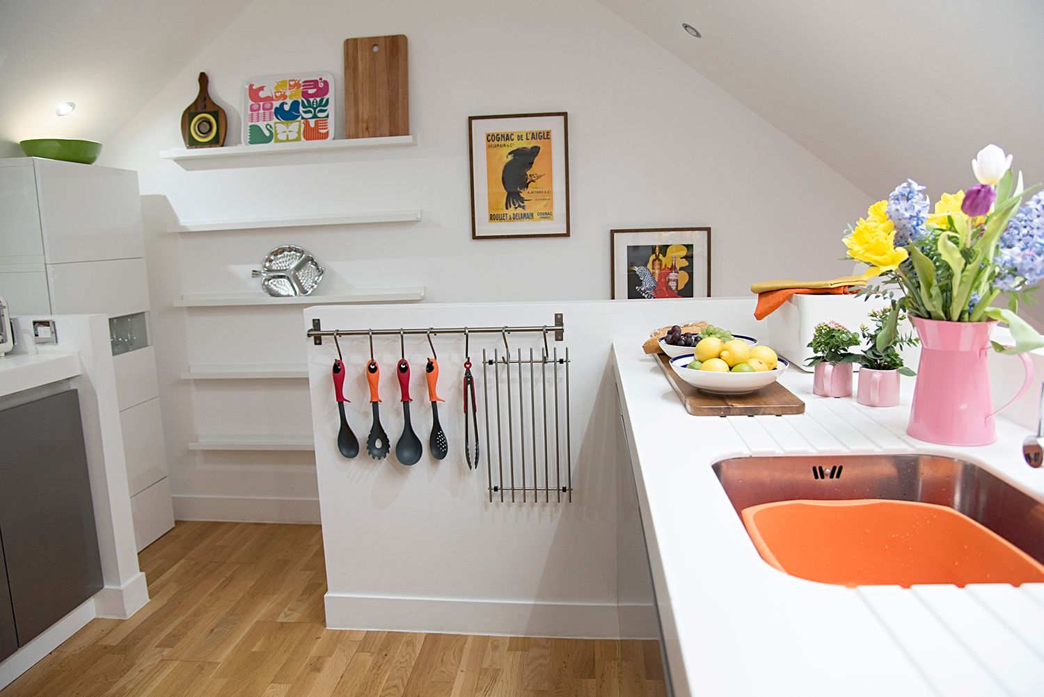 Modern white kitchen with sloped ceiling features floating shelves, colorful artwork, and spring flowers in a pink vase.