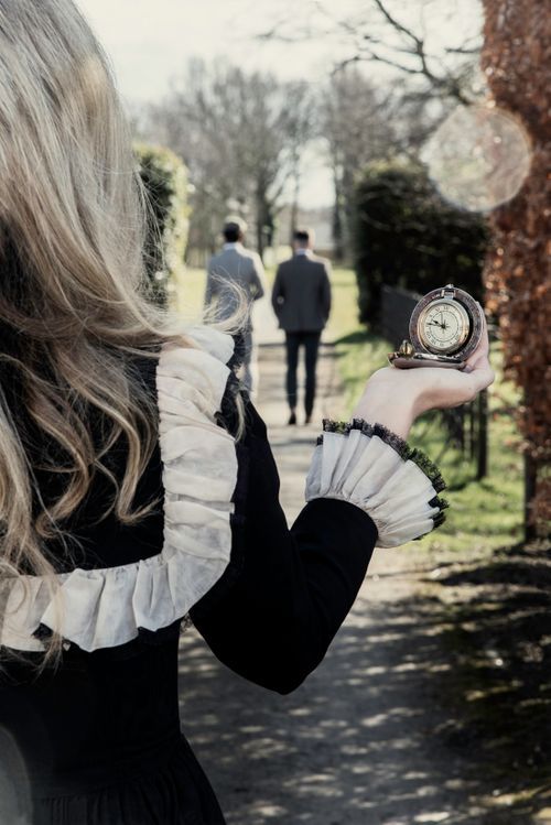 Woman in black dress holding vintage pocket watch as man walks away on park path.