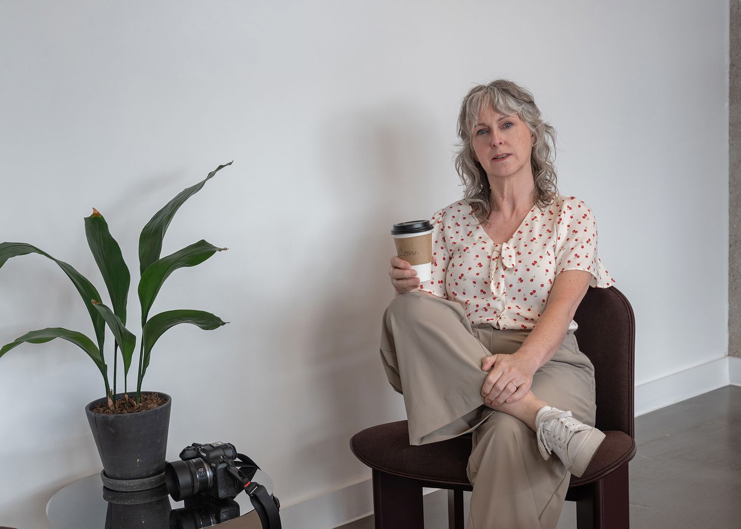 Woman with gray hair sits in chair holding coffee cup near a plant and camera in a bright white room.