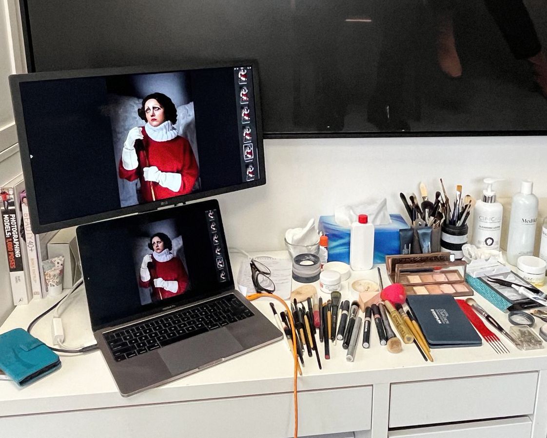 Photography workspace with laptop displays, makeup brushes, and styling tools on white desk.