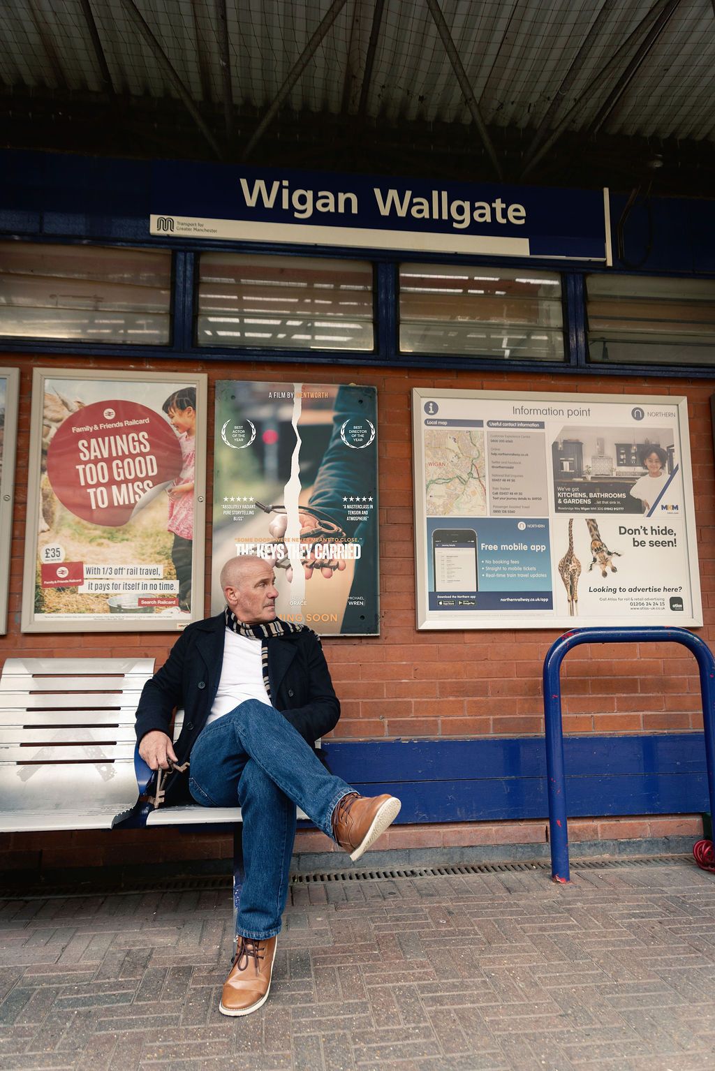 Man sitting on bench at Wigan Wallgate train station platform, relaxing beside advertisement boards.