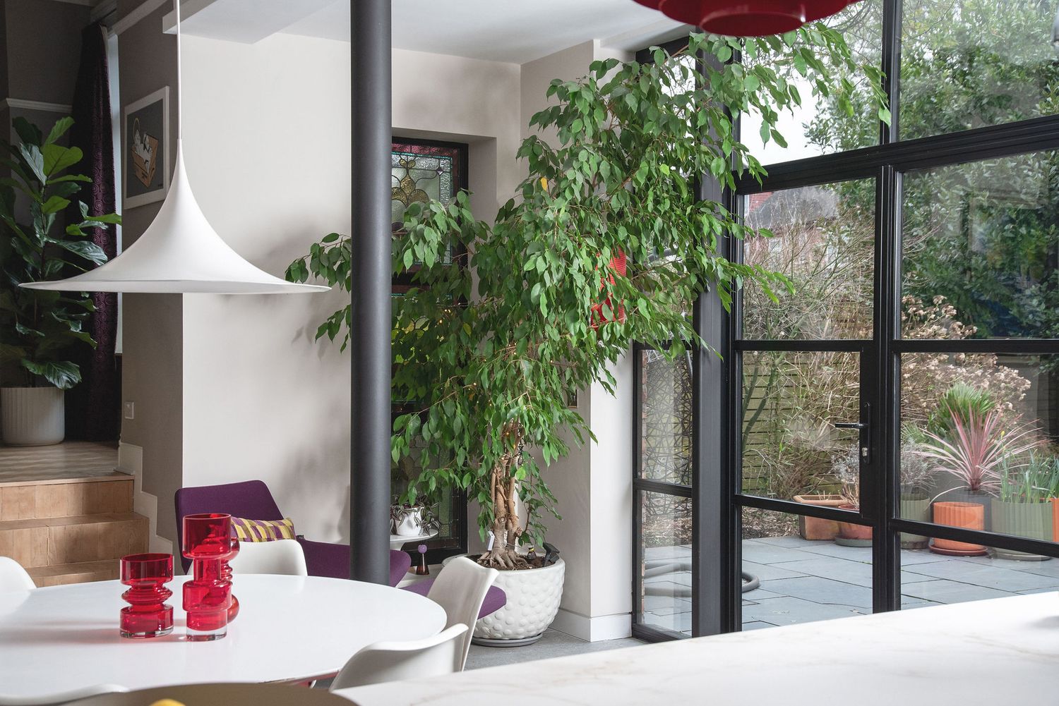 Modern interior space with large windows, indoor plants, and red pendant lamps creating a bright, natural atmosphere.