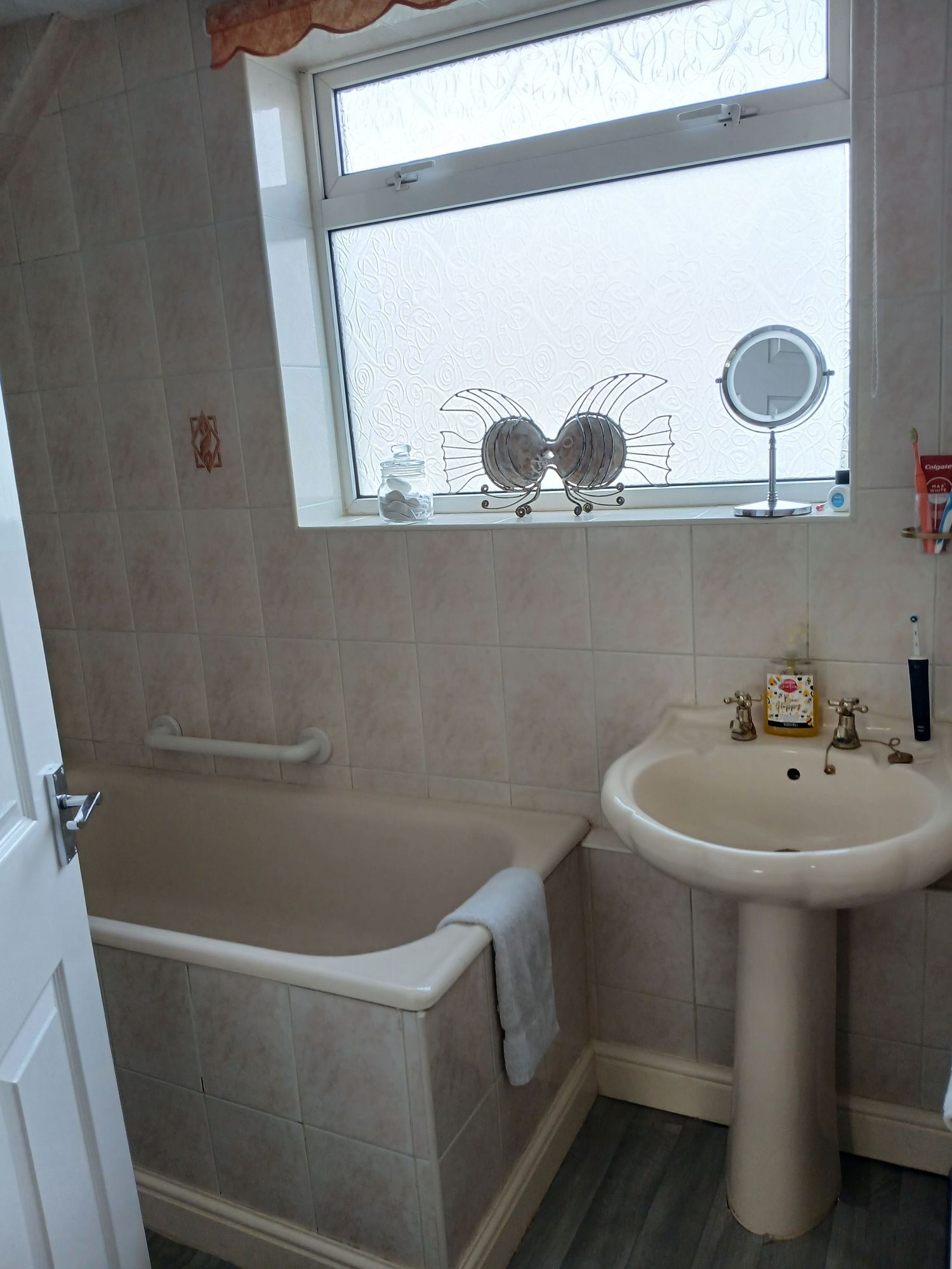 A modern white bathroom with gray tile walls features a pedestal sink, bathtub, and large window with decorative valance.