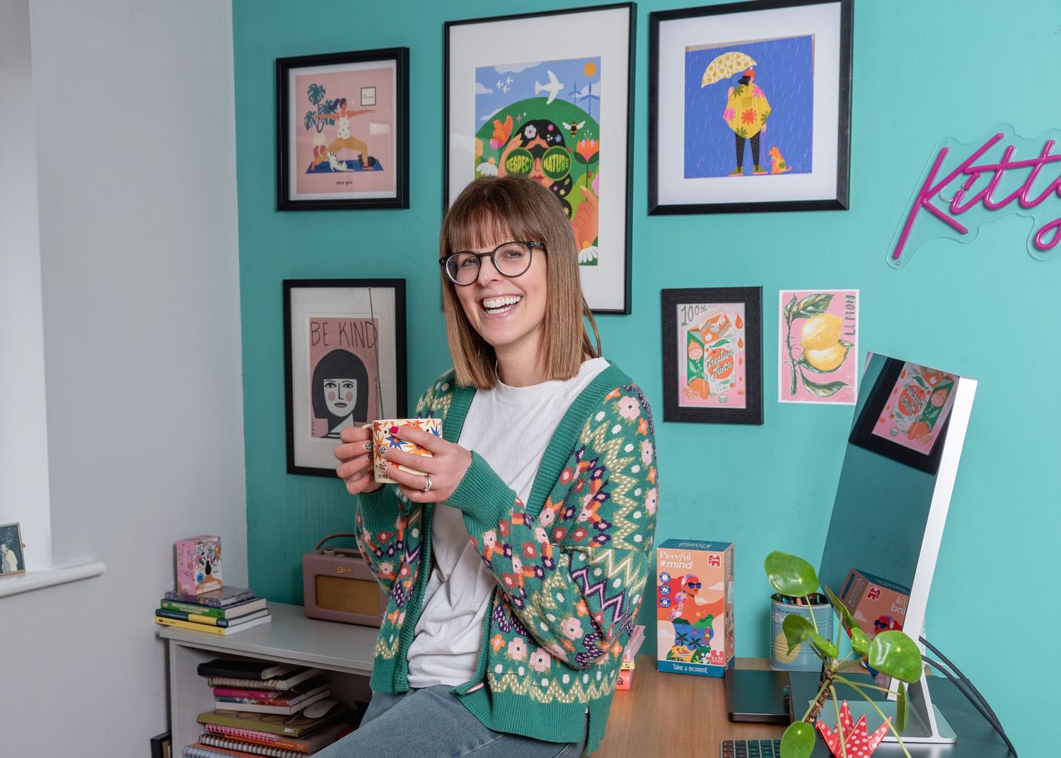 A person in glasses sits at a desk with teal walls decorated with framed artwork and illustrations.