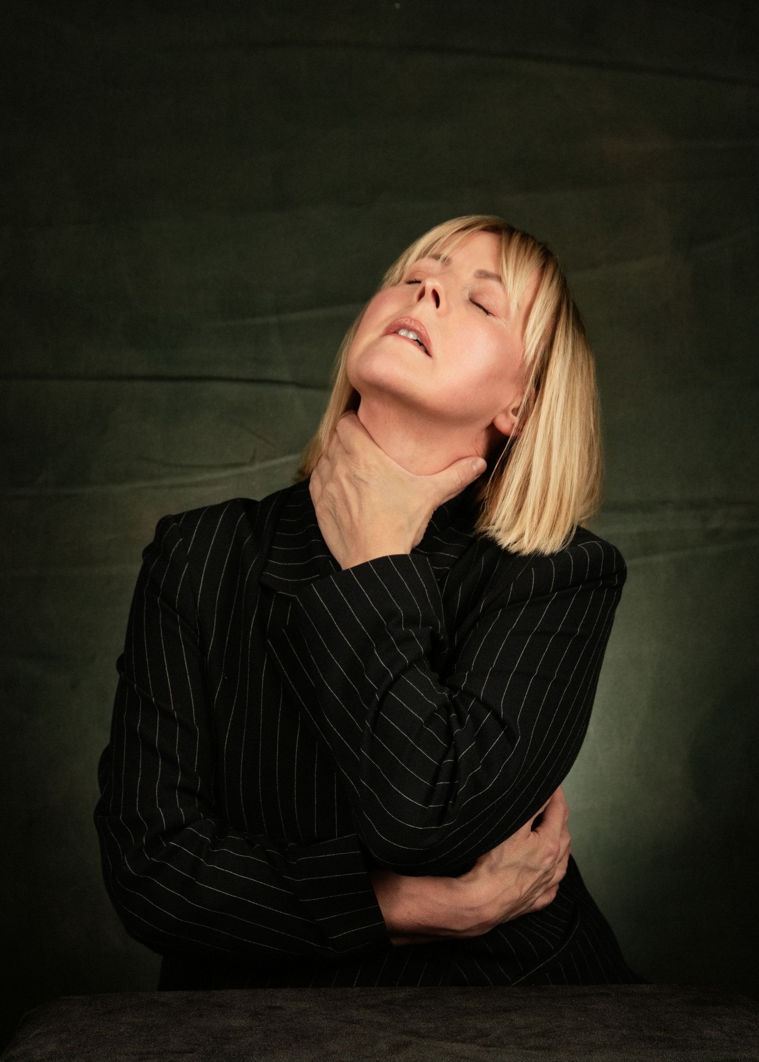 Blonde woman in black pinstripe blazer with head tilted back, eyes closed, hand on throat, dark background.