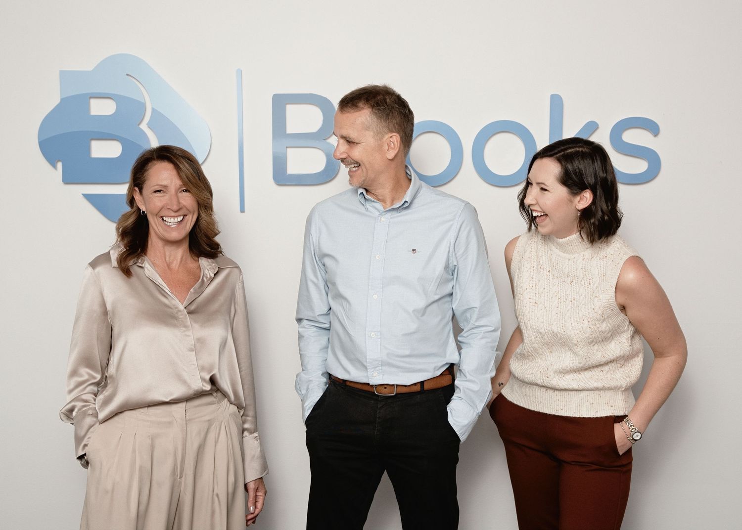 Three smiling professionals standing together in front of a Brooks company logo sign in a modern office.