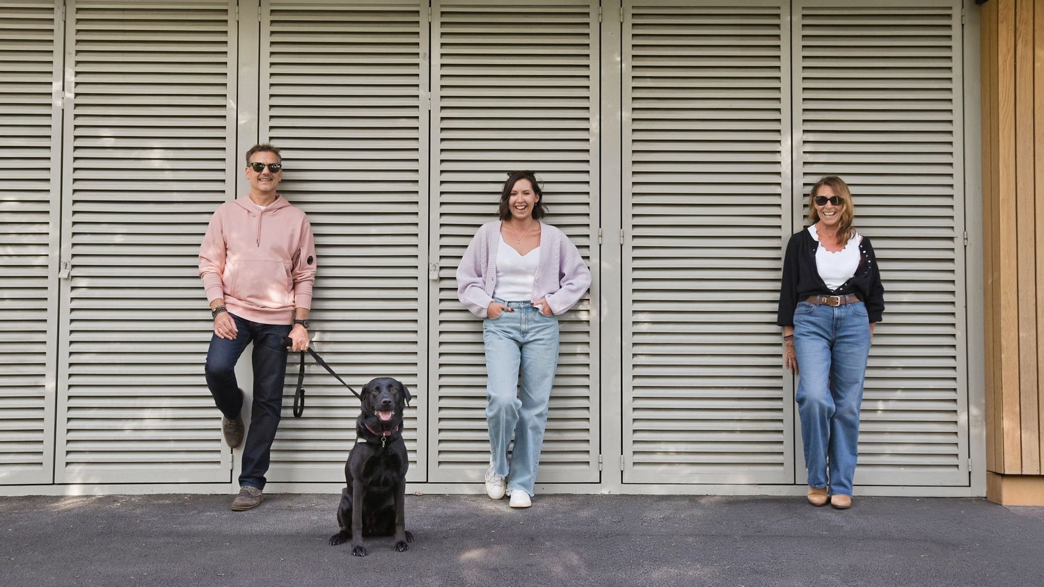 Three people and a black dog standing in front of a striped wall outdoors on a sunny day.