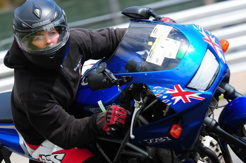 Motorcyclist wearing a black helmet rides a blue motorcycle decorated with a Union Jack flag sticker on a racetrack.