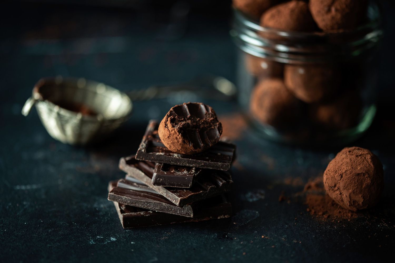 Dark chocolate squares stacked with cocoa-dusted truffles and a jar of chocolate truffles on a dark background.