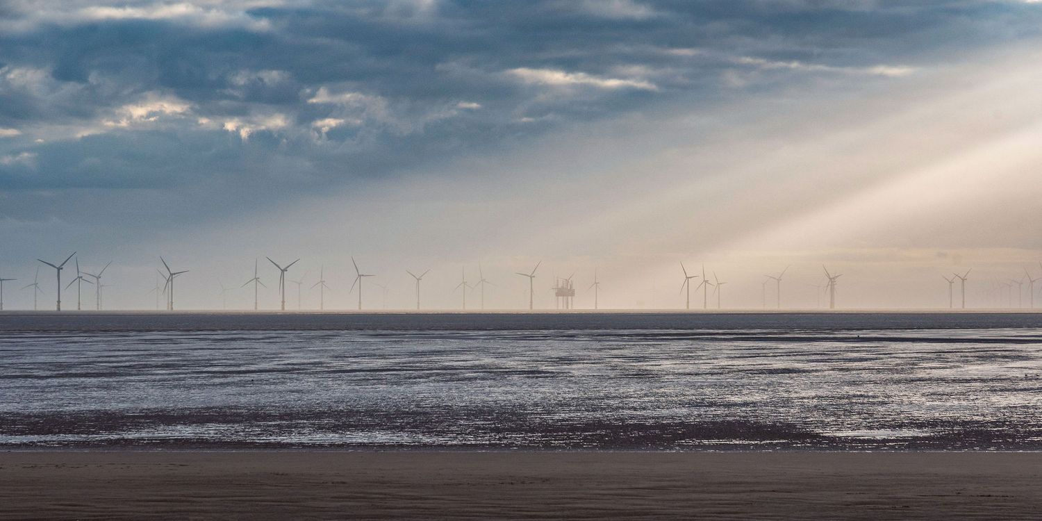 Offshore wind turbines stand in misty waters under dramatic sunbeams breaking through clouds over a sandy beach.