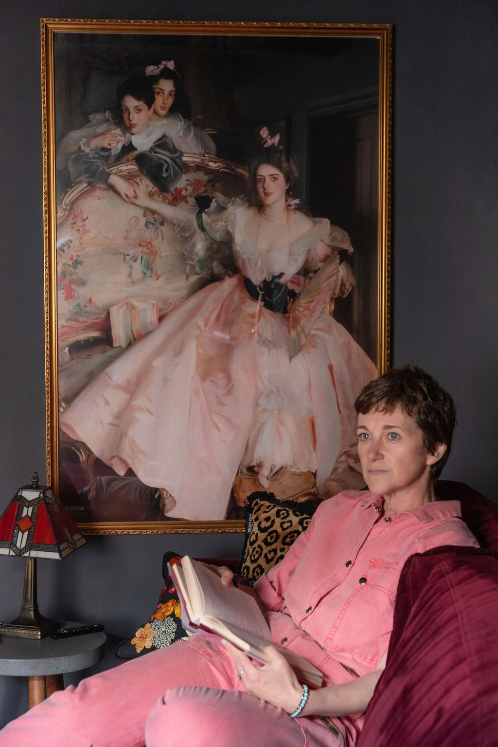 Woman in pink reads a book on a sofa beneath a classical portrait painting of elegant ladies in pink gowns.