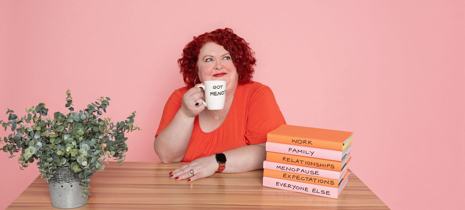 Woman with curly red hair in an orange top holds a white mug at a wooden desk with a stack of books and a potted plant against a pink wall.