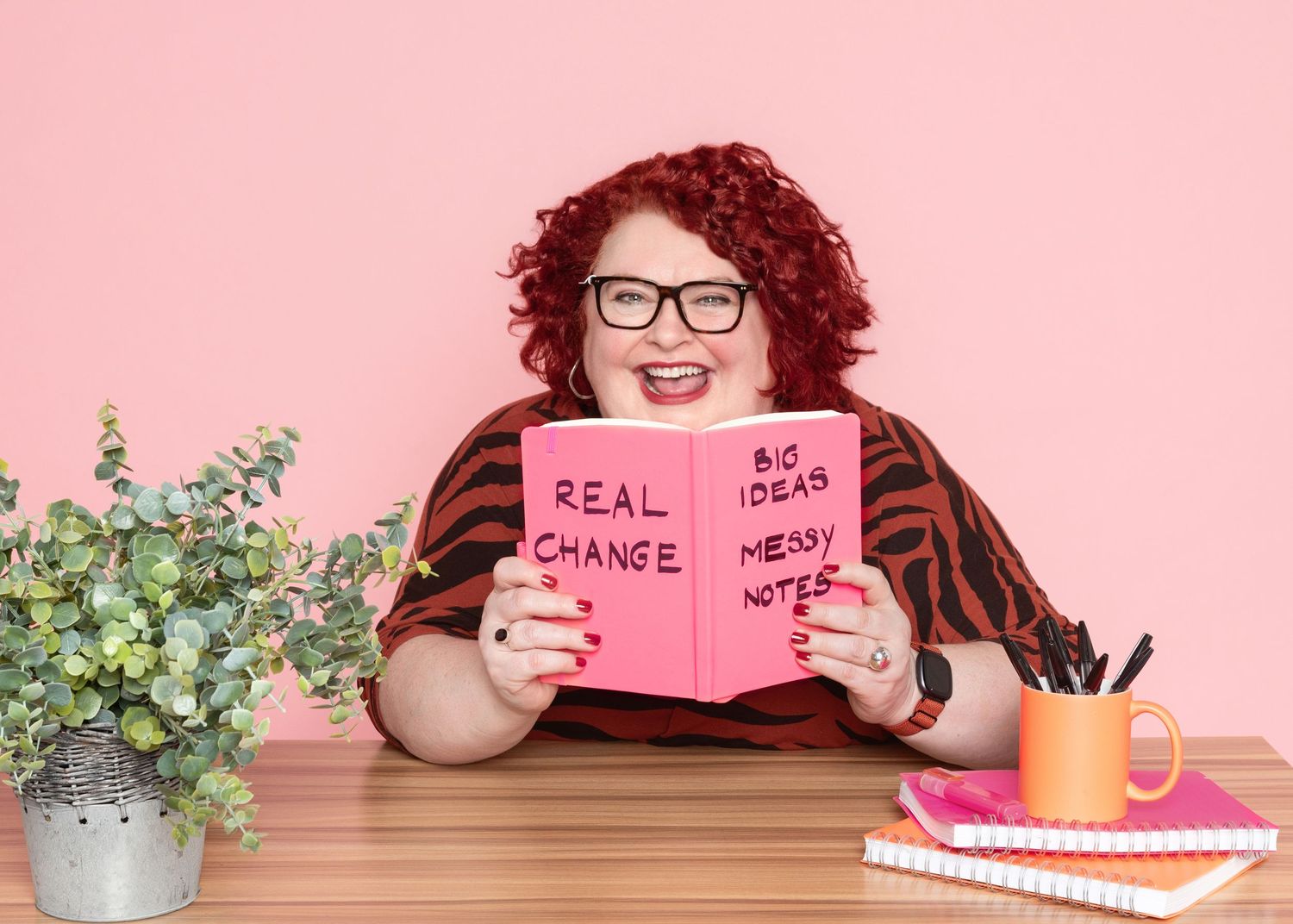 Smiling woman with red curly hair and glasses holds a pink notebook reading Real Change and Big Ideas at a desk with a plant and pens.