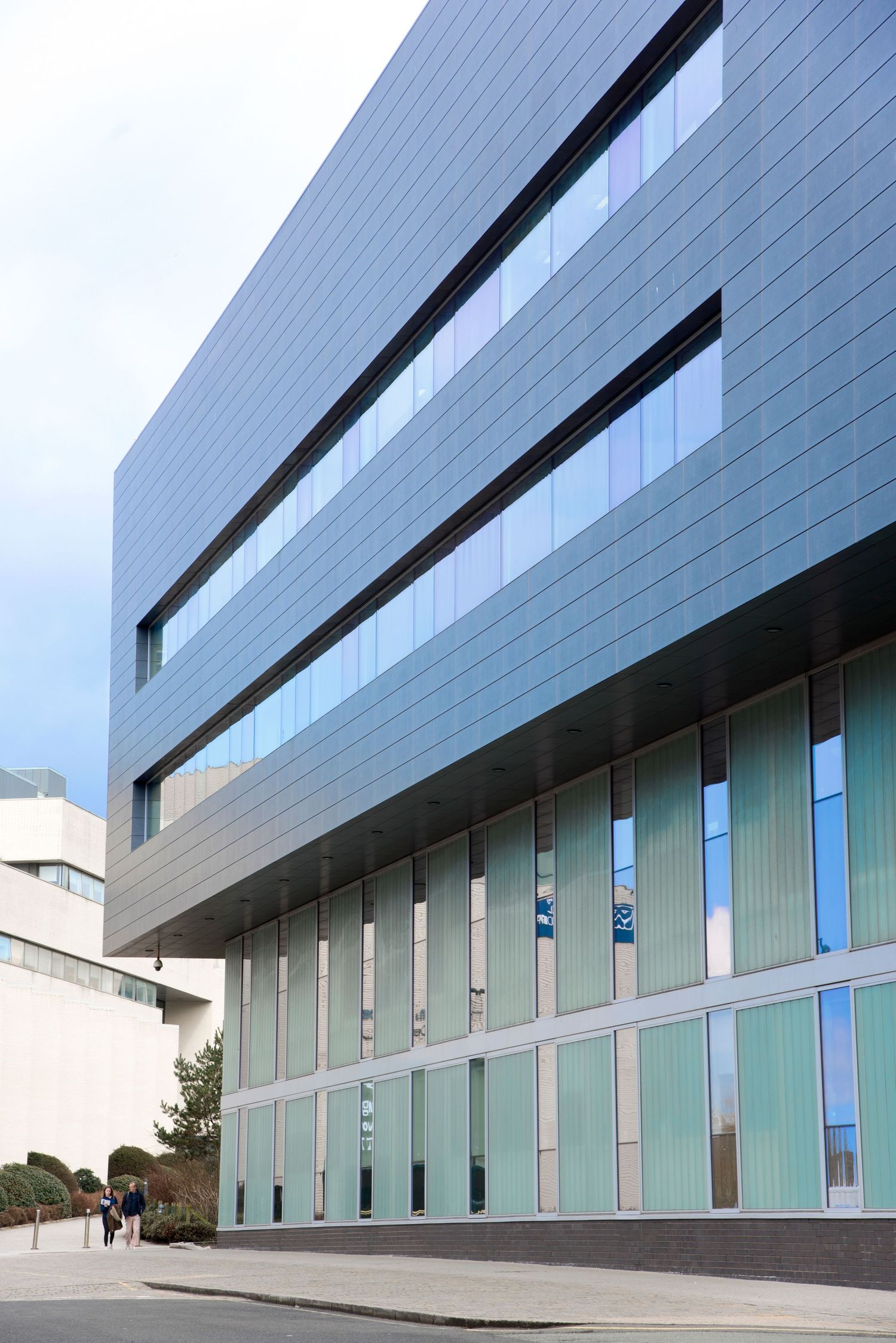 Modern glass and steel office building facade with reflective blue panels and pedestrians walking nearby.