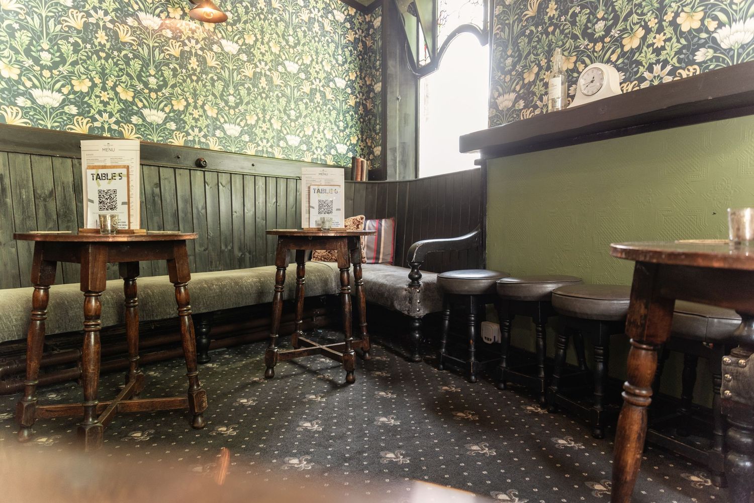Traditional British pub interior with ornate wallpaper, wooden bar stools, and tiled flooring in a cozy setting.