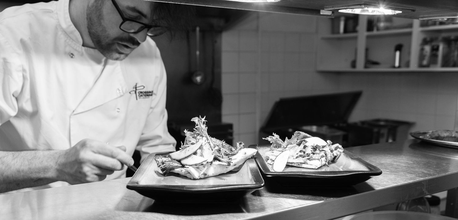 Chef in white uniform carefully plating gourmet dishes on dark plates in a professional restaurant kitchen.