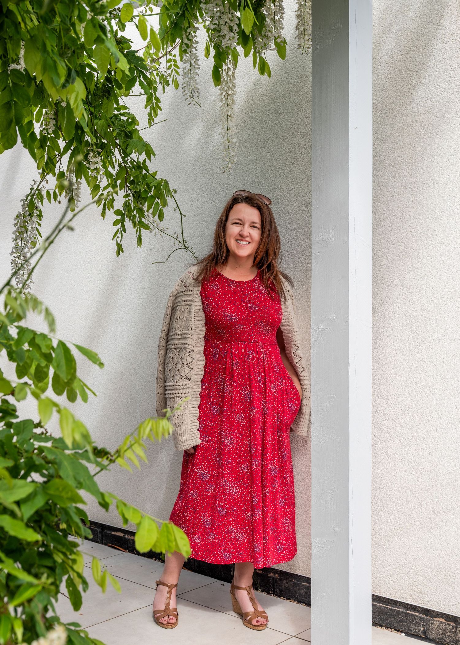 Woman in red floral dress and beige cardigan smiling outdoors near white wall with green foliage.
