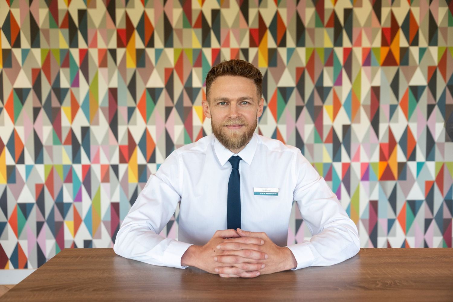 Professional businessman with beard sits at wooden desk before colorful geometric triangle pattern wall.