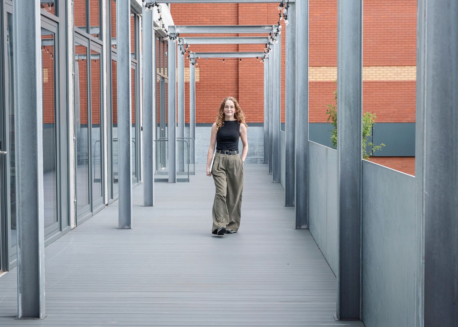 Woman in olive wide-leg pants and black top walking through modern glass corridor with brick building backdrop.