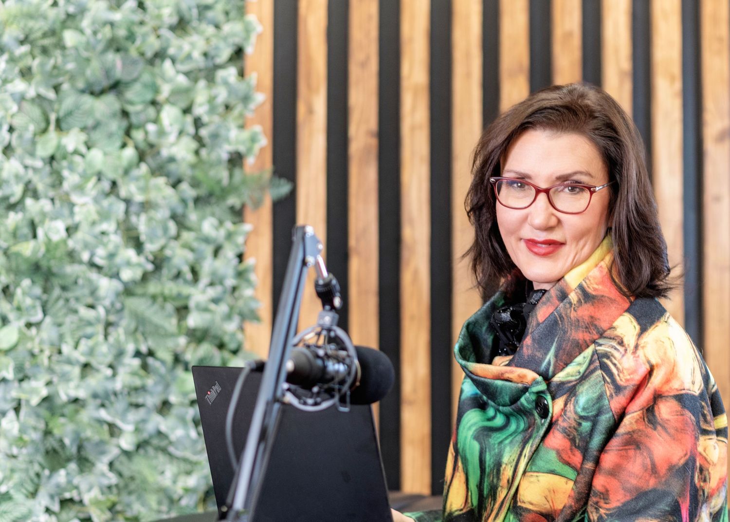A podcaster wearing red glasses and colorful clothing sits at a microphone with a green plant wall in the background.
