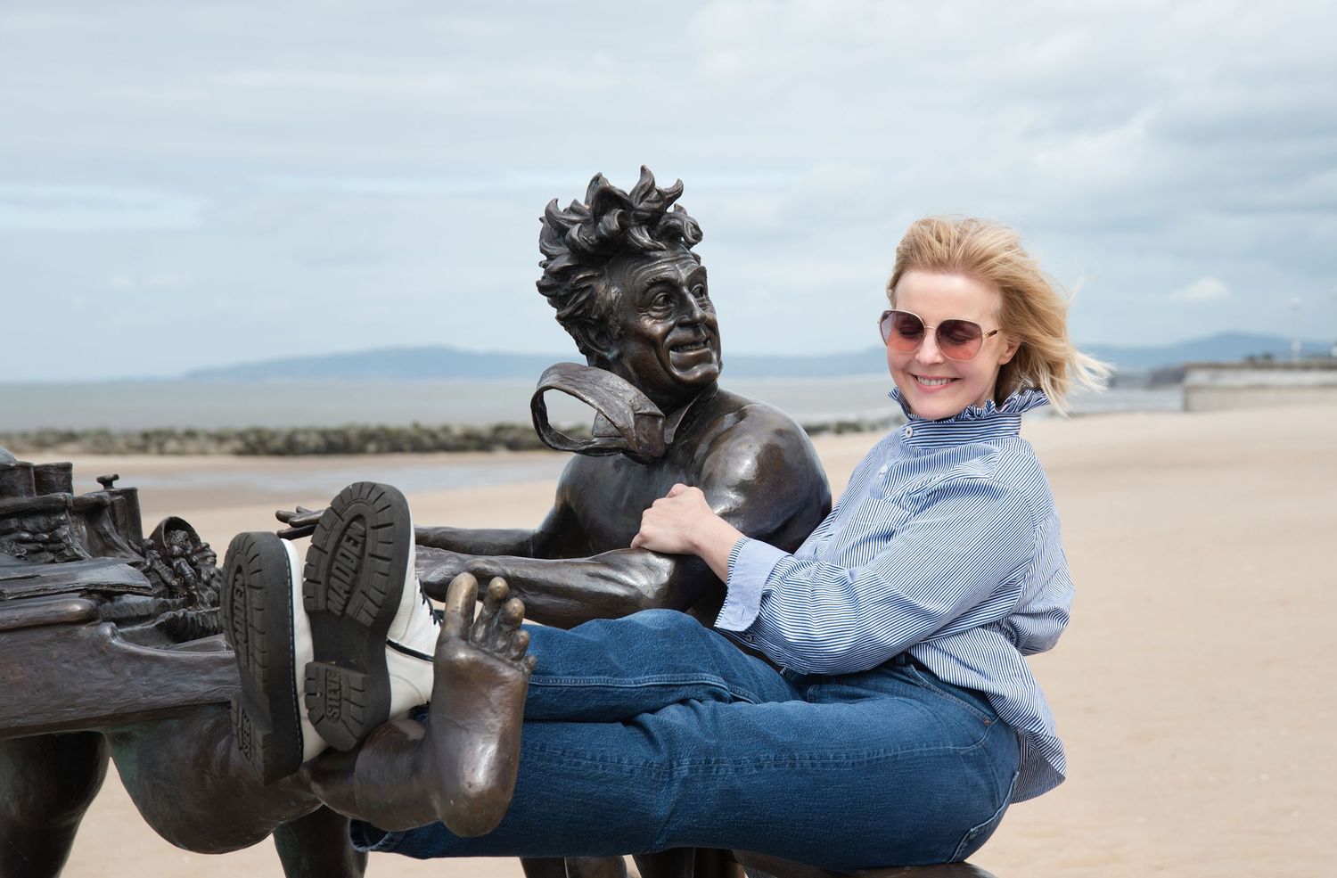 Woman posing playfully with a bronze statue on a sandy beach, mountains visible in the background.