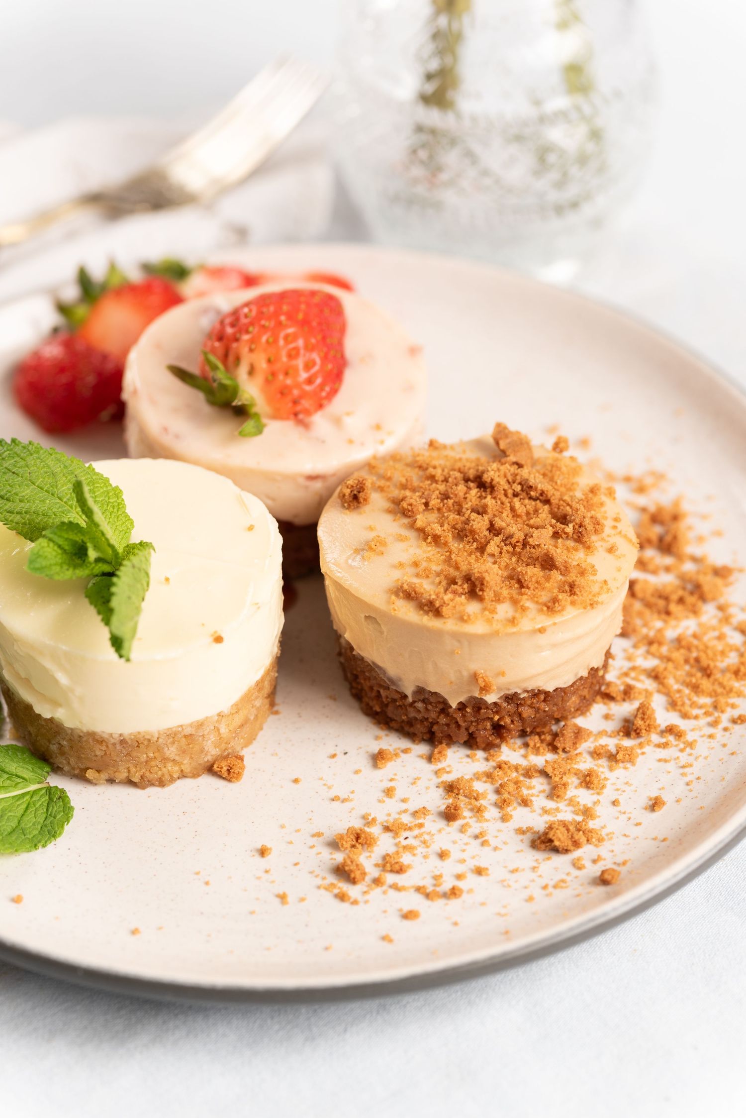 Three mini cheesecakes on a plate, topped with strawberries, mint, and cookie crumble on a white surface.