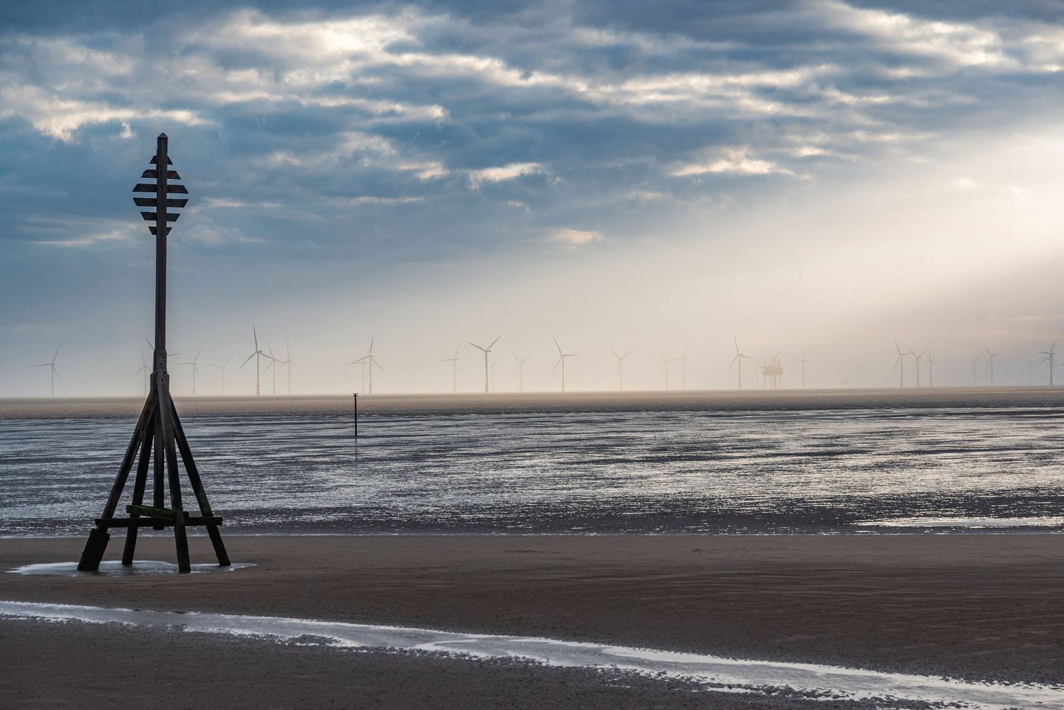 Beach scene with a navigational marker, wet sand, and offshore wind turbines under dramatic cloudy skies.