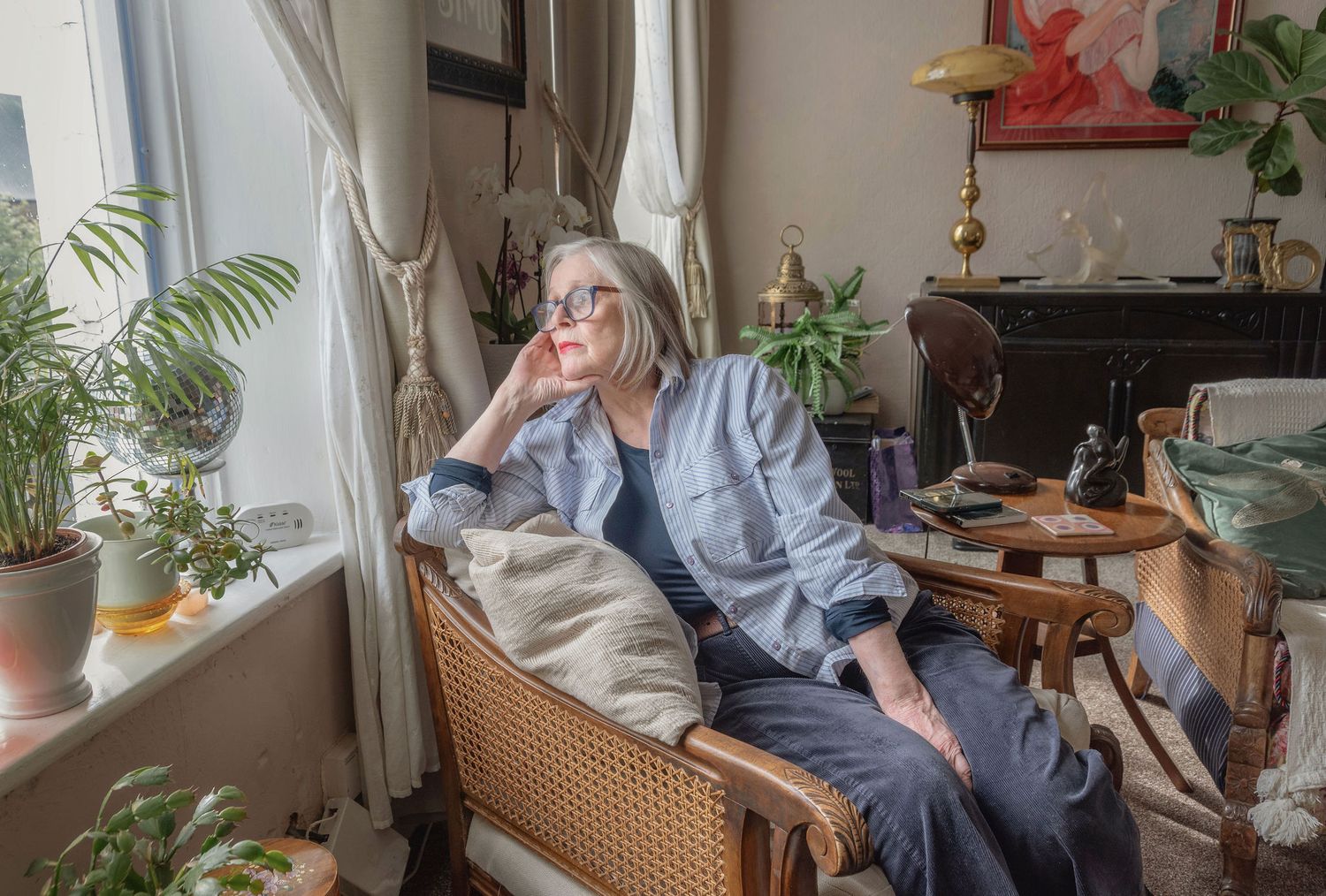 Older woman with gray hair and glasses sits thoughtfully in a wicker chair in a cozy, plant-filled living room.