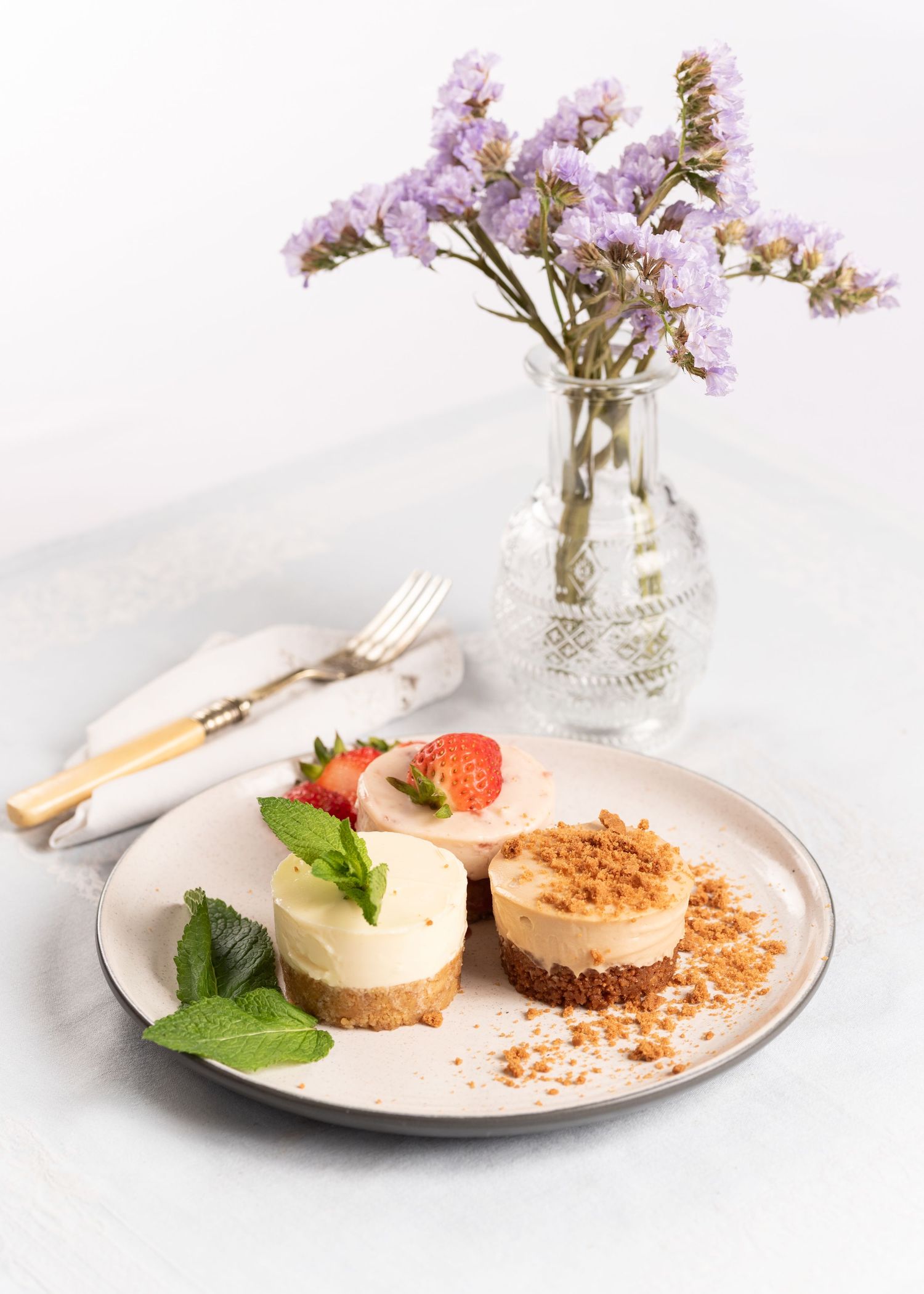 Two mini cheesecakes on a plate with strawberries and mint, beside a vase of purple flowers and cutlery.