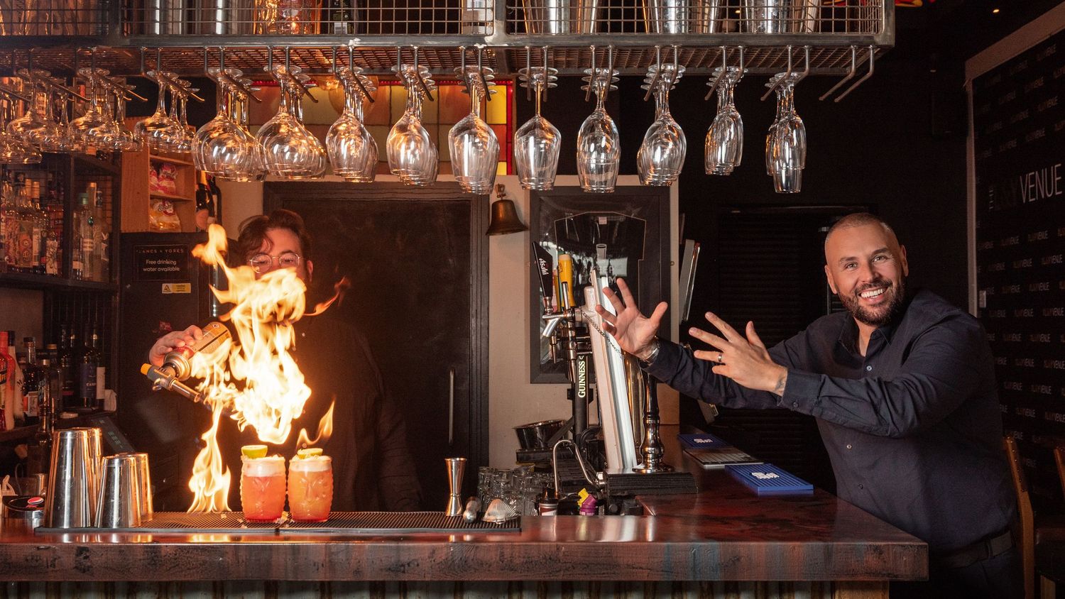 Smiling bartender gestures beside a flaming cocktail preparation at a stylish bar with wine glasses hanging overhead.