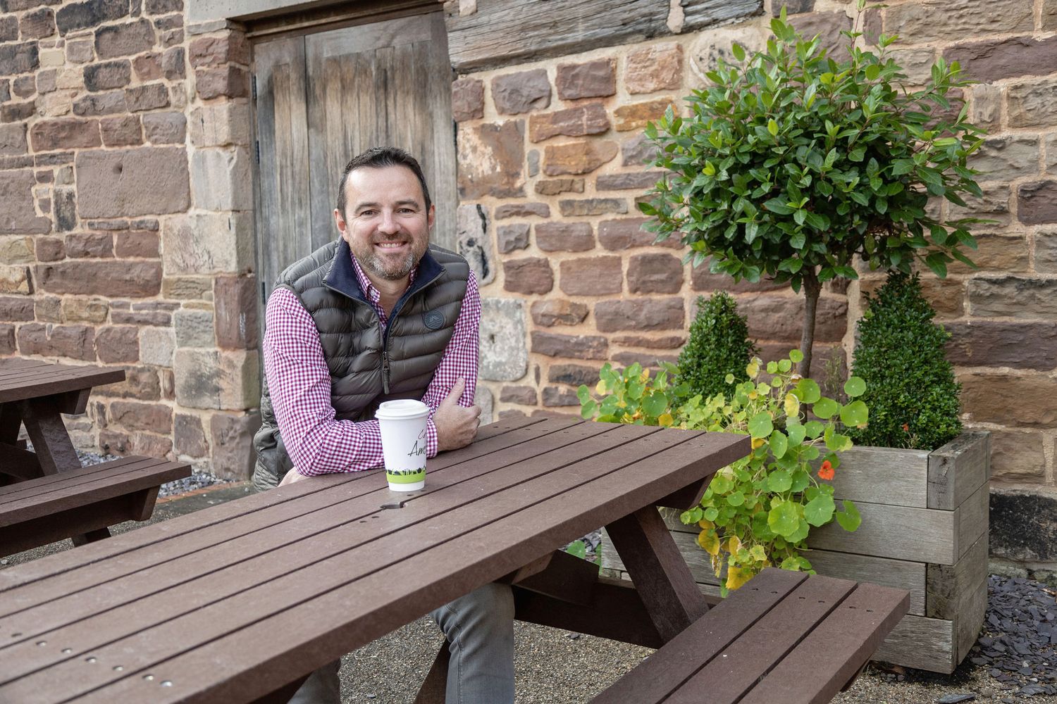 Man smiling at outdoor picnic table with coffee cup beside stone wall and decorative topiary plants.