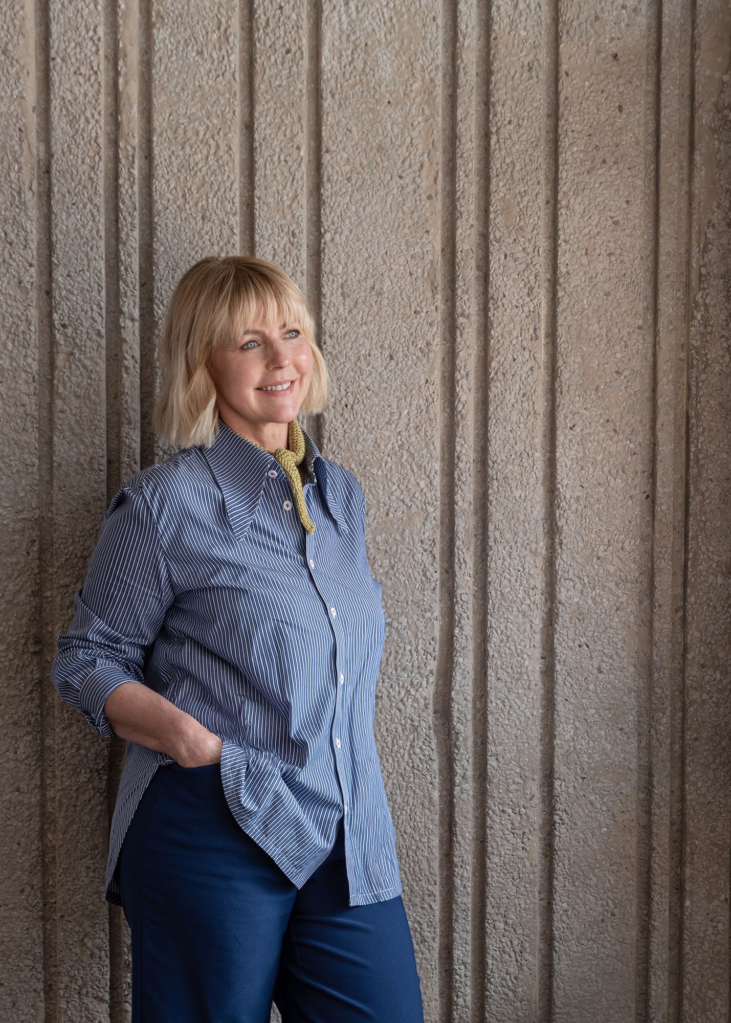 Smiling blonde woman in blue striped shirt and teal pants leaning against a textured concrete wall.