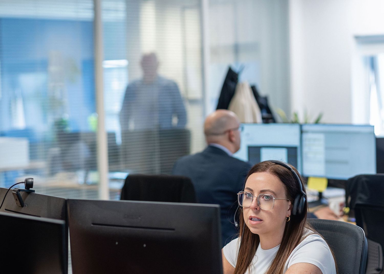 Woman with headset and glasses works at computer in modern office, colleagues visible through glass partition in background.