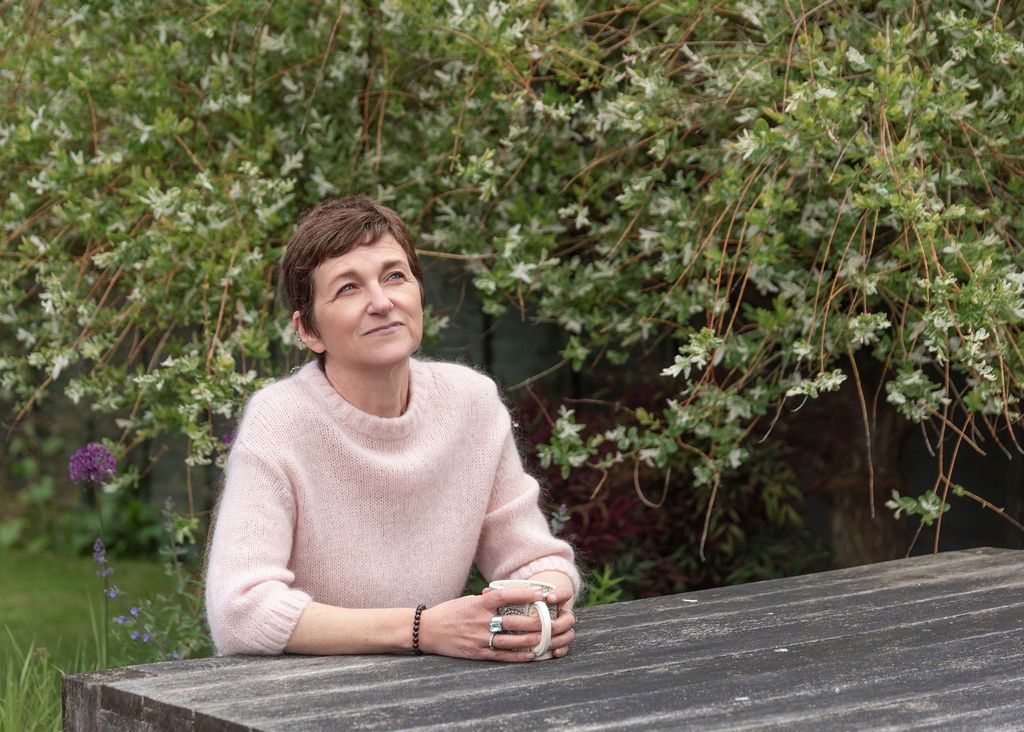 Woman in pink sweater sitting at outdoor wooden table, smiling thoughtfully in a lush green garden setting.