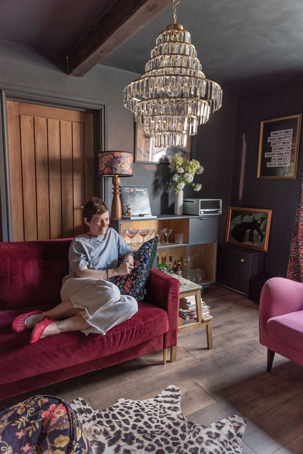 Woman relaxing on red velvet sofa beneath crystal chandelier in eclectic dark-walled living room with animal print rugs.