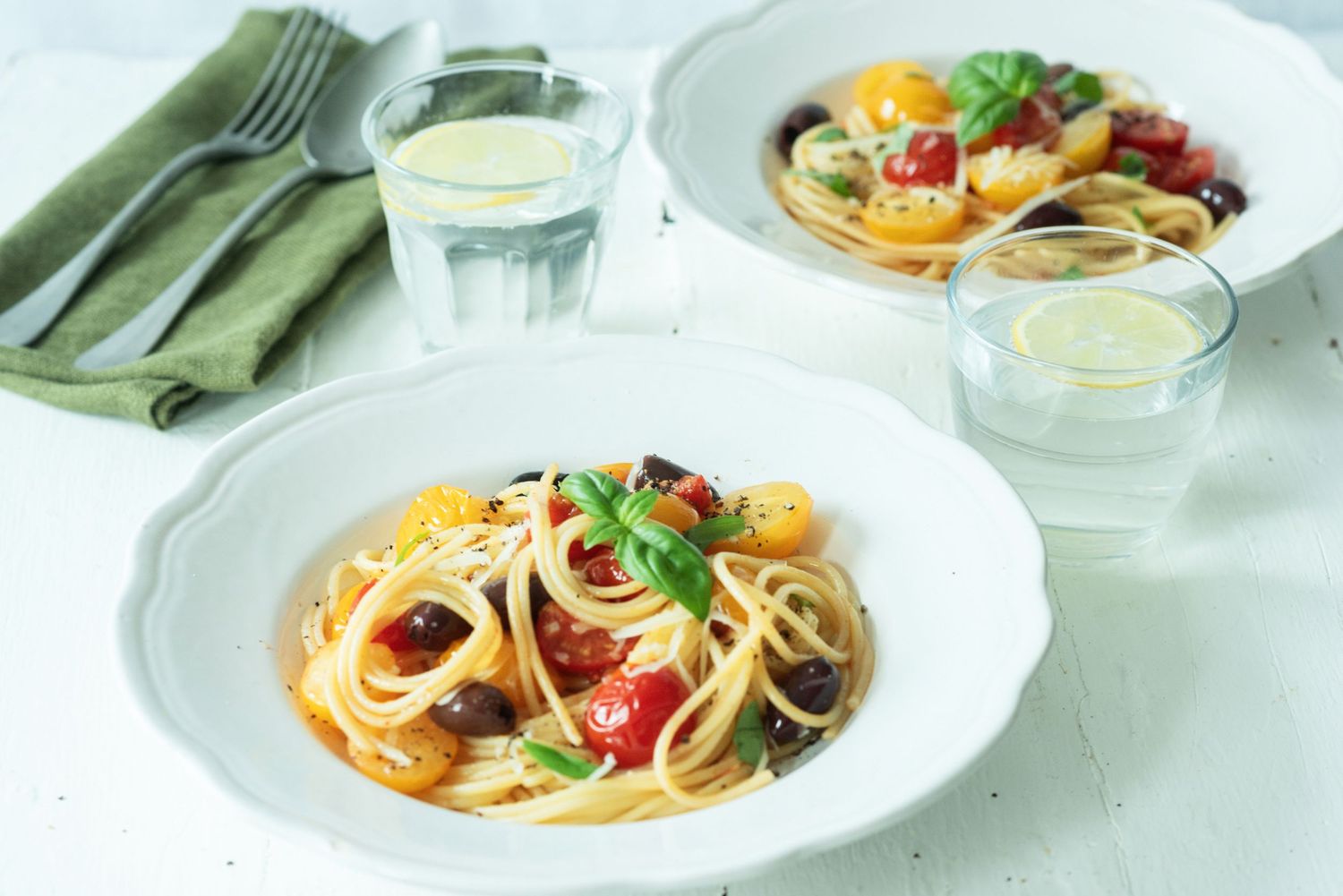 Two white bowls of spaghetti with cherry tomatoes, olives, and basil, served with glasses of water on a white table.