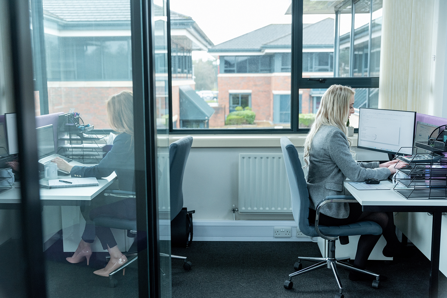 Office worker at desk with computer monitors beside window in modern workspace.